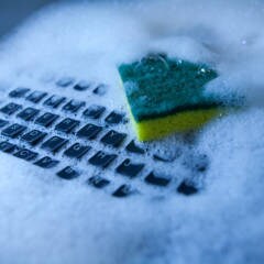 A computer keyboard being cleaned in soap suds and water with a yellow kitchen sponge.